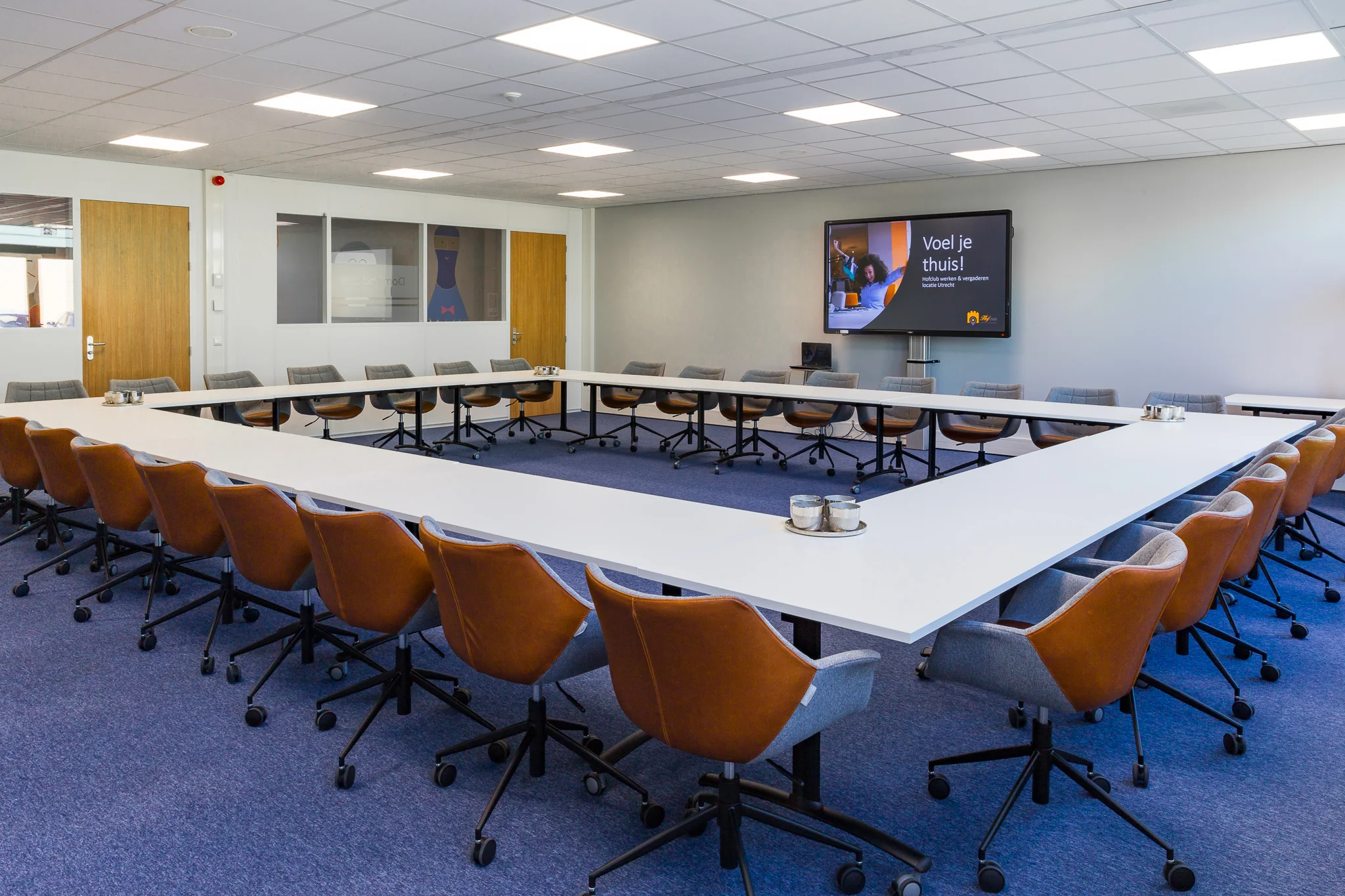 Vergaderruimte aan de Niels Bohrweg met een U-opstelling van witte tafels, bruine en grijze bureaustoelen en een presentatiescherm aan de muur.